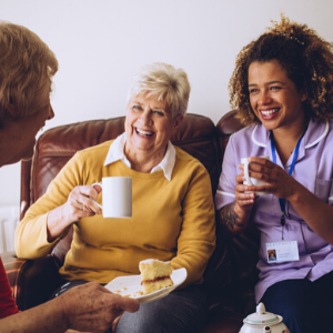 Elderly carer sitting with two of her patients in the care home. They are enjoying some cake and tea.